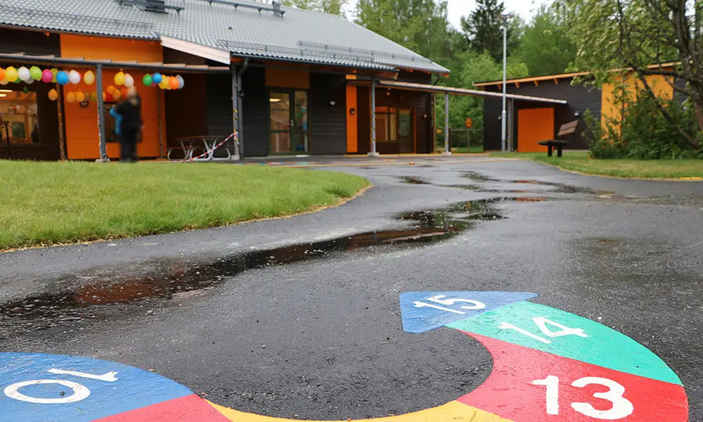 Foto på en del av Lärlingens förskola. Det är en grå och orangefärgad envåningsbyggnad. På asfalten på förskolans gård finns en färgglad pil med siffror i. 